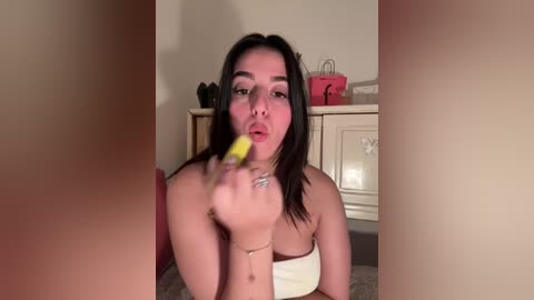 Media: Video of a young woman with dark hair and medium skin tone, wearing a strapless white top, eating a yellow banana, in a cozy, dimly lit kitchen with beige cabinets.