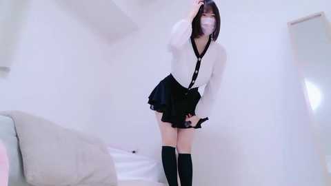 Media: Video of an Asian woman in a white blouse, black pleated skirt, and knee-high socks, wearing a face mask, standing in a minimalist white room with a gray couch.