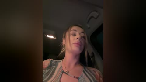 Media: Video of a fair-skinned woman with long brown hair, wearing a sheer, patterned top, leaning forward in a dimly lit car interior.