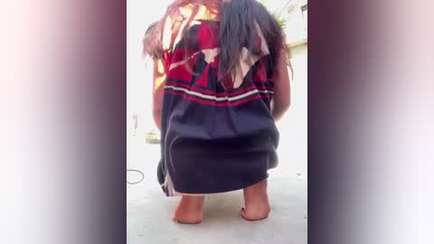 Media: Video of a person with long dark hair, wearing a traditional, colorful, patterned skirt and barefoot, standing on a light-colored surface. Background shows blurred outdoor elements.