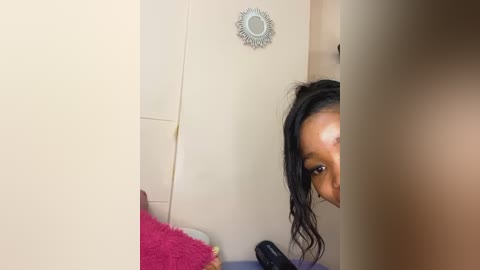 Media: A video of a young Black woman with dark skin, curly hair, and a neutral expression, partially hidden by a beige wall and a decorative round mirror. The background features a pink pillow and a black phone on a bed.