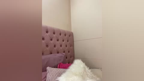 Media: A video of a cozy bedroom corner featuring a tufted, pink upholstered headboard, a fluffy white throw blanket, and a soft, lavender pillow. The background is a plain, cream-colored wall.