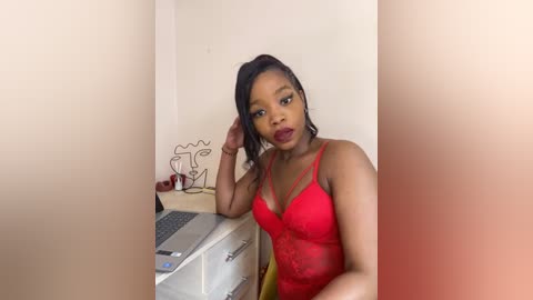 Media: Video of a Black woman with medium skin tone and long, dark hair in a high ponytail, wearing a red lace bra, leaning on a white desk in a minimalist room.