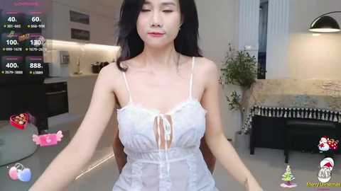 Media: Video of a young East Asian woman with long black hair, wearing a white lace lingerie bodysuit, standing in a modern kitchen with white cabinets and black appliances.