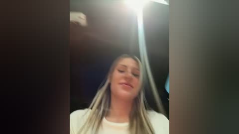 Media: Video of a young, fair-skinned woman with long, straight blonde hair, smiling slightly, wearing a white t-shirt, standing in a dimly lit room with a blurry, indistinct background.