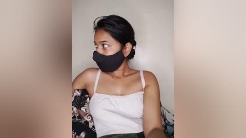 Media: Video of a young South Asian woman with medium skin tone, dark hair, and a black mask. She wears a white tank top and black arm sleeves with floral patterns. The background is a plain beige wall.