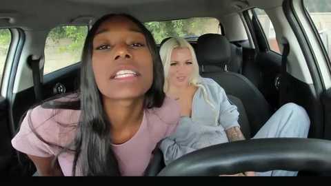 Media: A candid video inside a car with two women; a dark-skinned woman in pink, and a light-skinned blonde in blue, both smiling.