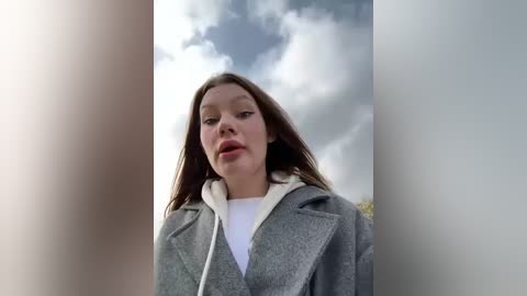 Media: Video of a young Caucasian woman with long brown hair, wearing a grey coat over a white t-shirt, looking upward at a cloudy sky.