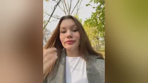 Media: A video of a young woman with long brown hair, wearing a white shirt and gray blazer, standing outdoors in a park with trees and green foliage. The image has a soft focus and is divided into three sections with different colors.