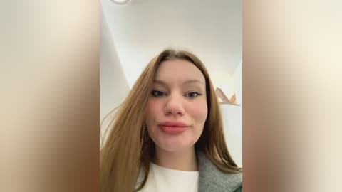 Media: Video of a young Caucasian woman with long, straight brown hair, fair skin, and a neutral expression, wearing a grey jacket and white shirt. The background features a white wall and a blurred, indistinct object.