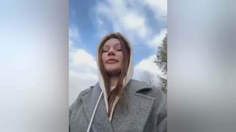 Media: Video of a Caucasian woman with straight, light brown hair, wearing a grey hooded coat, standing against a cloudy blue sky backdrop, looking serene.