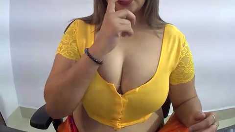 Media: Video of a curvy woman with light brown skin, wearing a yellow lace-trimmed crop top that accentuates her large breasts. She sits on a black chair with orange fabric.