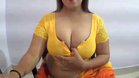 Media: Video of a woman with medium skin tone, long dark hair, wearing a yellow lace-trimmed top that is unbuttoned to expose cleavage, sitting on a couch with a bright orange cloth draped over her lap.