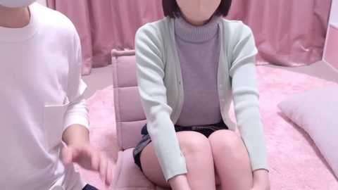 Media: Video of a young East Asian woman with shoulder-length black hair, wearing a light green cardigan over a lavender turtleneck, sitting on a pink quilted bed. A man in a white shirt stands beside her. Pink curtains and walls surround the scene.