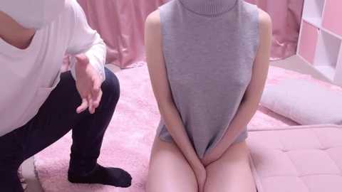 Media: Video of a slender, light-skinned woman with a small bust in a grey turtleneck, sitting on a pink tufted bed, with a man in a white shirt and black pants standing behind her, touching her arm.