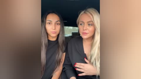 Media: Video of two women, one with long, straight, dark hair, the other with long, straight, blonde hair, both in black blazers, sitting closely together in a car.