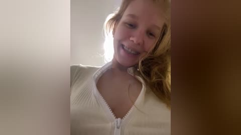 Media: Video of a joyful young girl with fair skin, blonde hair, and a broad smile, wearing a white zip-up sweater, set against a softly blurred background.