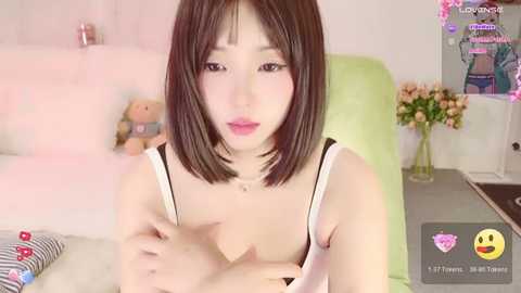 Media: A video of an East Asian woman with straight black hair and fair skin, wearing a white camisole, sitting on a bed with pink and green decor.