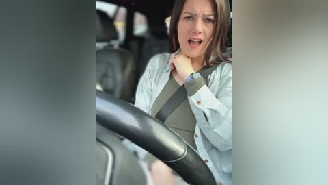 Media: Video of a distressed woman in a car, wearing a light jacket and green top, gripping the steering wheel, mouth open in distress, blurred car interior background.