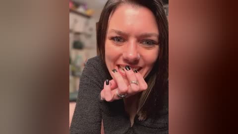 Media: Video of a Caucasian woman with straight brown hair, wearing a gray sweater, biting her nails with black-painted nails and a silver ring. Background features a blurred kitchen with cabinets and shelves.