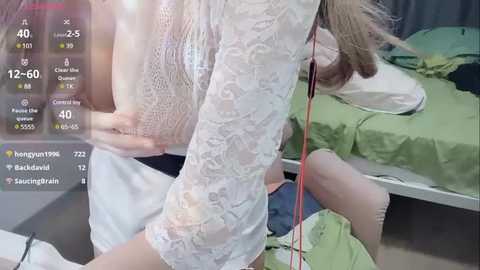 Media: A video of a woman with light skin, wearing a white lace dress, standing next to a hospital bed with green sheets. Her left hand is on her stomach, and she appears to be in a hospital room.