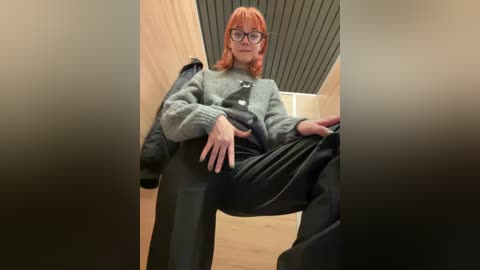 Media: Video of a person with red hair, glasses, and a gray knit sweater, sitting on a black chair in a modern room with wooden walls and a ceiling.