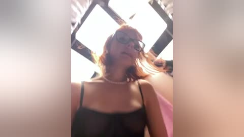 Media: A video of a fair-skinned woman with shoulder-length red hair, wearing black-framed glasses, a black spaghetti strap top, and a pearl necklace, looking up through a triangular window.