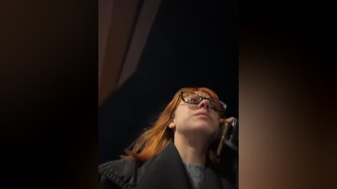 Media: Video of a woman with red hair, wearing black-rimmed glasses and a black blazer, looking up intently, against a dark, blurred background.