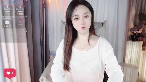 Media: A video of an Asian woman with long, straight black hair, fair skin, and wearing a white long-sleeved shirt, standing indoors with a white radiator behind her.