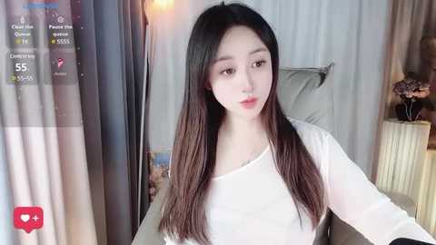 Media: Video of an East Asian woman with long, straight black hair, fair skin, and a slender figure, wearing a white long-sleeved top, seated indoors with a white curtain and a radiator in the background.