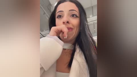 Media: A candid video of a young woman with long black hair, wearing a white choker and white blouse, biting her index finger, taken indoors with blurred background.