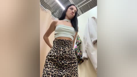 Media: Video of a young woman with long black hair in a leopard print skirt, wearing a white strapless top, standing in a beige-walled changing room with a white coat and floral jacket hanging nearby.