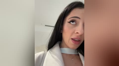 Media: Video of a woman with long black hair, wearing a white choker, and white blazer, with a distressed expression, against a plain white wall.