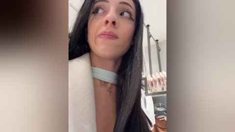 Media: Video of a young woman with long, straight black hair, fair skin, and a choker necklace, looking up with a contemplative expression. Background shows a room with a metal rack and shelves.