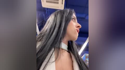 Media: Video of a woman with long, straight black hair, wearing a white choker and a gray jacket, seen in profile against a blurred, dark background with an air conditioning unit visible.