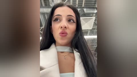 Media: Video of a young woman with long black hair, fair skin, and full lips puckered in a kiss, wearing a white coat and light blue top, in a modern industrial setting with metallic ceiling and pipes.