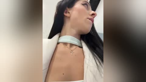 Media: Video of a fair-skinned woman with long black hair, wearing a light blue choker necklace, and a white jacket, seen from the side in a blurred indoor setting.