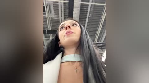 Media: Video of a young woman with long dark hair, wearing a white top and a choker necklace, looking upward in an industrial setting with exposed metal beams.