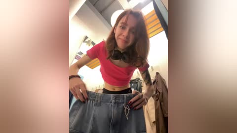 Media: Video of a young woman with fair skin and shoulder-length brown hair, wearing a pink crop top, black bra, and blue high-waisted shorts, standing in a dimly lit room with a window and hanging clothes in the background.