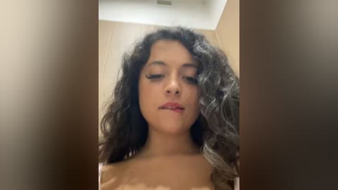 Media: Video of a young woman with long, curly black hair, light brown skin, and full lips, captured from a low angle, showing her upper chest.