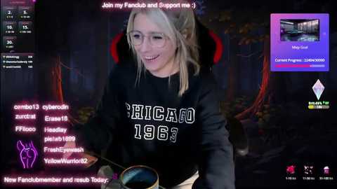 Media: Video of a young blonde woman with glasses, wearing a black \"Chicago 1866\" sweatshirt, smiling while playing a video game. The background features a dark, dungeon-like environment with red and purple lighting.