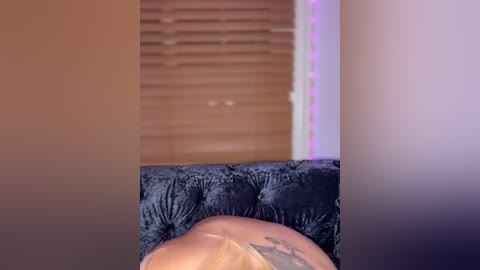 Media: A video of a woman's bare back and tattoo, seen from behind, reclining on a tufted velvet chair. The background features horizontal blinds and purple LED strip lights.