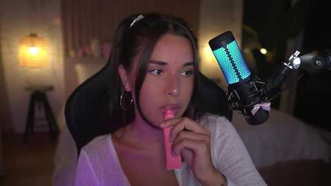 Media: Video of a young woman with medium skin tone and long dark hair, wearing a light gray top, holding a pink lollipop near a green microphone. Background shows a dimly lit room with a bed, lamp, and bookshelf.