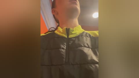 Media: Video of a person wearing a neon yellow and gray puffer jacket, slightly blurred with a warm orange hue. The background shows a dimly lit room with a circular ceiling light.