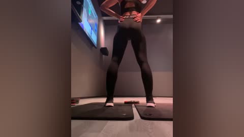 Media: Video of a woman in tight black leggings and sports bra, standing on exercise mats in a dimly lit gym, with a TV screen displaying a blue and white image in the background.