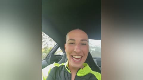 Media: Video of a smiling woman with light skin and short hair, wearing a neon green and black jacket, inside a car. The background shows blurred foliage and sky.