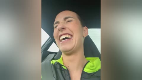 Media: Video of a young man with short hair, wearing a black and neon green jacket, laughing with a wide smile in a car, blurry background.