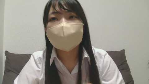 Media: Video of an Asian woman with long black hair, wearing a white shirt, beige face mask, and brown pillow behind her.