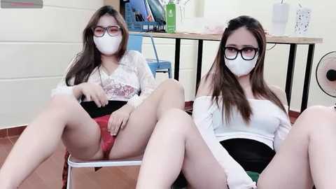 Media: Video of two Asian women in surgical masks, white lab coats, and black skirts, sitting on chairs, legs spread, revealing red lace panties. Background includes a desk, computer, and fan.