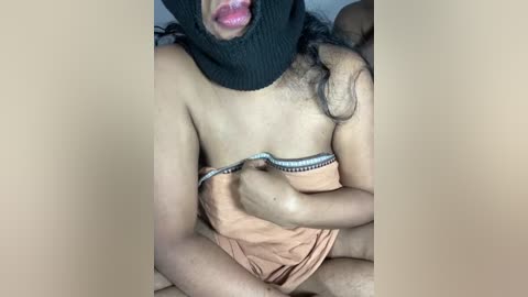 Media: Video of a person with a dark knit cap, partially covering their face, and wearing a beige towel wrapped around their torso, exposing their bare chest and part of their arm.